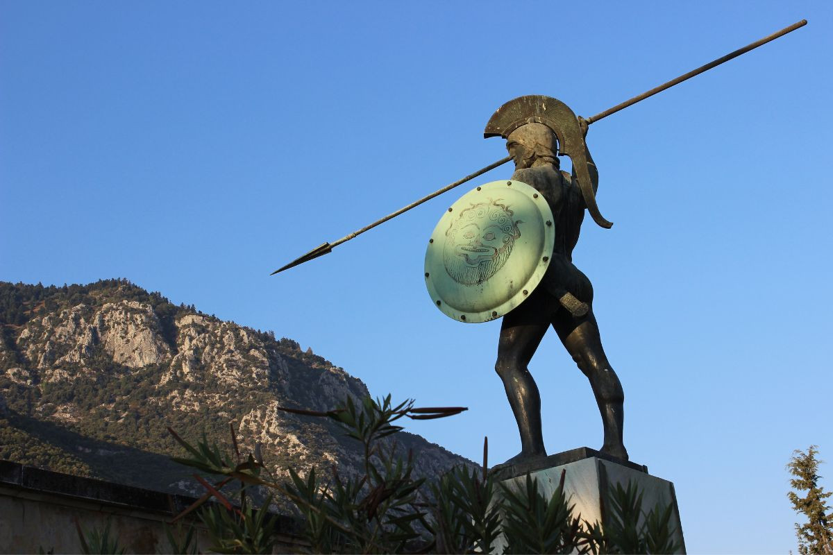 A statue of a Greek warrior holding a spear and shield at Thermopylae, backed by a rocky hillside.